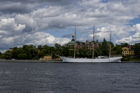 White three-masted sailing ship docked in Stockholm harbor with historic buildings and dramatic cloudy sky